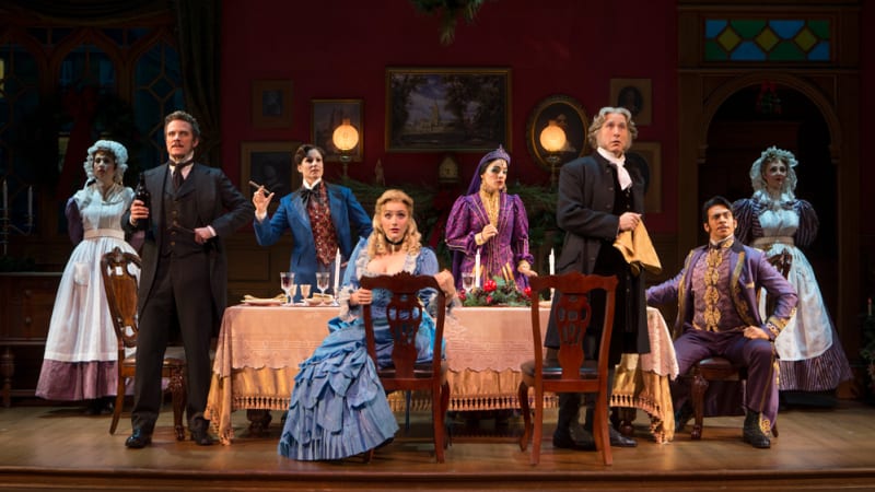 A group of actors in fancy 1870s clothes stand and sit around a dining table with two maids in the background.