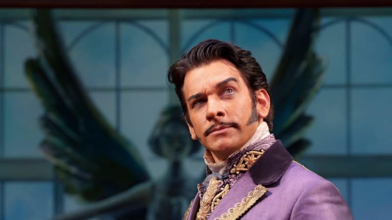 A man with dark hair and sharp sideburns wears a long purple coat with fancy gold accents. He raises an eyebrow in suspicion.