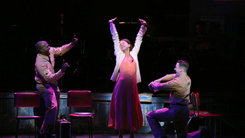 A woman in a peach dress and white jacket raises her arms up in glee, and two soldiers in tan uniforms cheer her on.