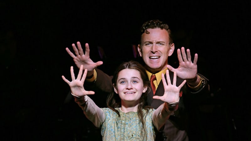 A young girl in a green dress holds her hands out in front of her with her fingers spread. A man in a brown suit stands behind her and does the same pose.