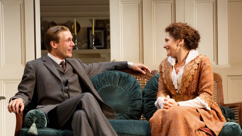 A man dressed in a gray 1900s suit and a woman in an orange 1900s gown sit on a cushioned bench and speak to each other.