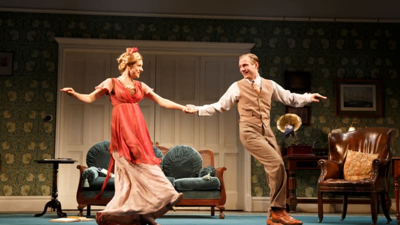 A blonde woman and man in early 1900s clothing dance together in a living room with green patterned wallpaper.