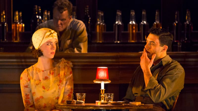 A woman in a yellow floral dress and 1930s hat sits at a small table at a bar with a man who smokes a cigarette.