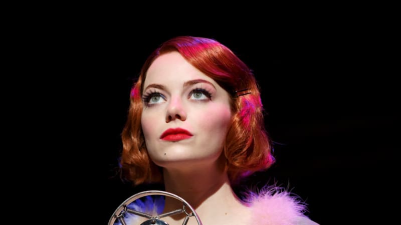 A woman in a short red wavy bob stands at a 1920s microphone. She wears a slip dress under a mint green lavish robe with fur and fringe