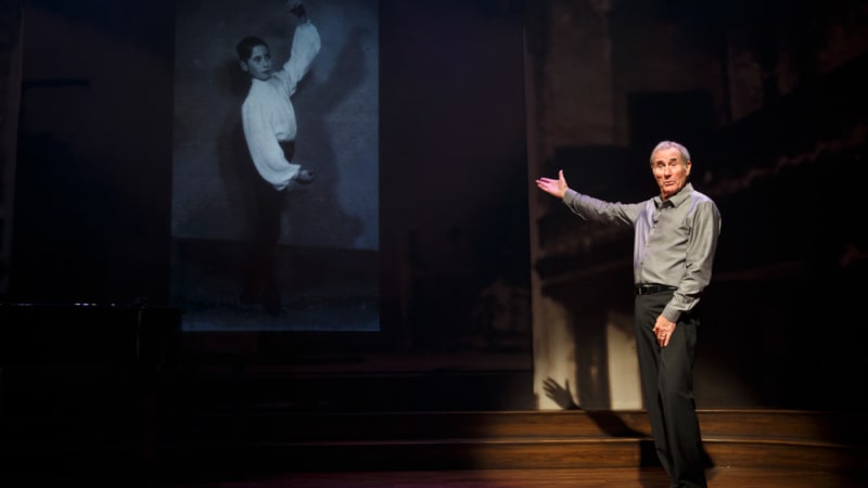 A man with gray hair wearing a gray collared shirt gestures to a projection of a black and white photo of a young boy in a dance pose.