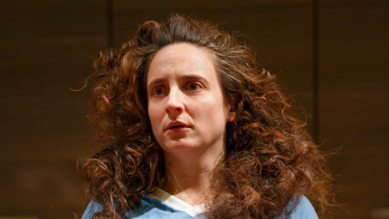 A woman with long, messy brown curls clutches her stomach with concern. She wears a causal big shirt and sweatpants.