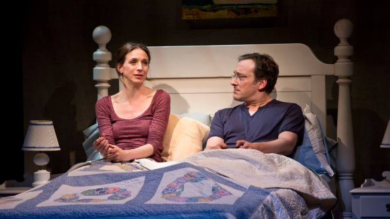 A woman and man sit up together in bed, resting against a headboard and covered by a quilt. They both look a bit concerned.