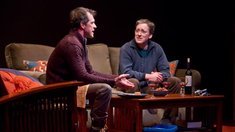 Two men in zip up sweaters sit on a sofa and have a tough conversation. On the coffee table in front of them are a few glasses of wine and some food.