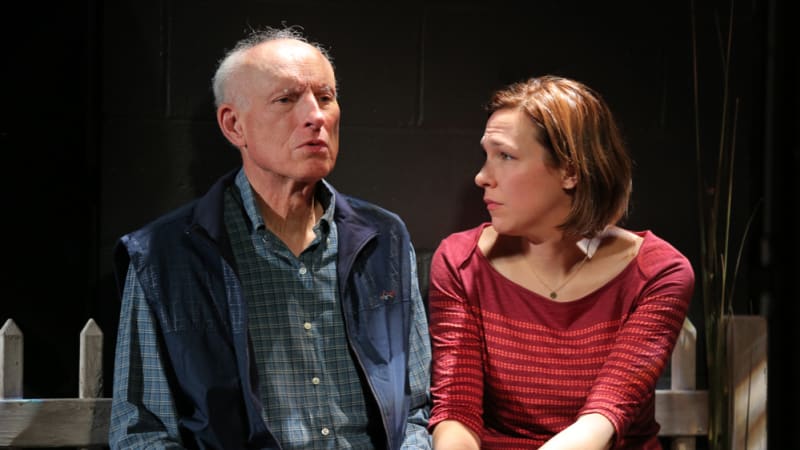 A woman sits next to a young man and holds his hand. She wears a dark red striped shirt and has short hair. He wears a blue plaid shirt under a collared vest.