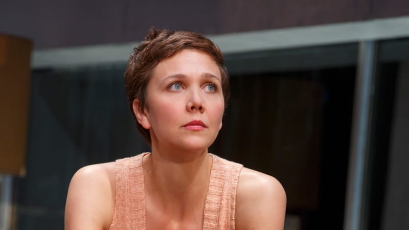 A woman with bright blue eyes and a short brown pixie cut sits on a turquoise softa and holds a bowl with a spoon in it. She wears a light peach tank top and a floral skirt.