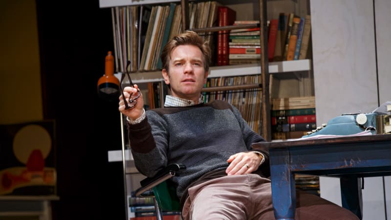 A man with reddish brown hair leans back in a desk chair and holds his glasses in one hand. He wears a collared shirt under a sweater and khakies.