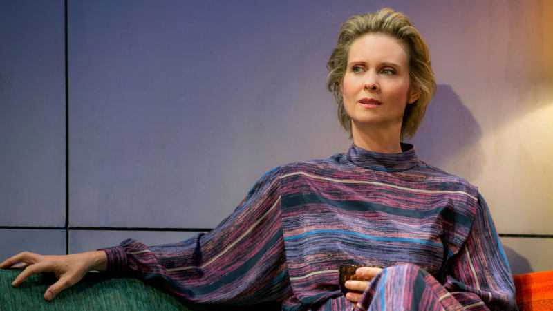 A woman with short blonde hair wears a multi-colored striped dress and sits on a turquoise softa with a drink in hand.