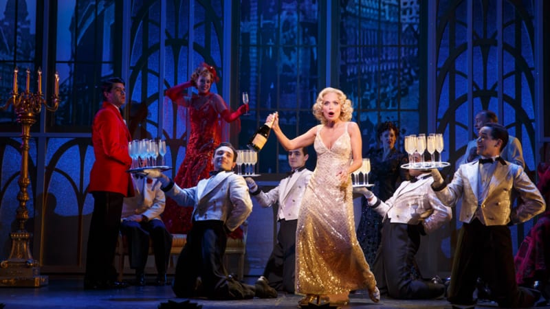A blonde woman in a sparkly dress holds a bottle of champagne. Behind her are four waiters dancing with trays of champagne glasses.