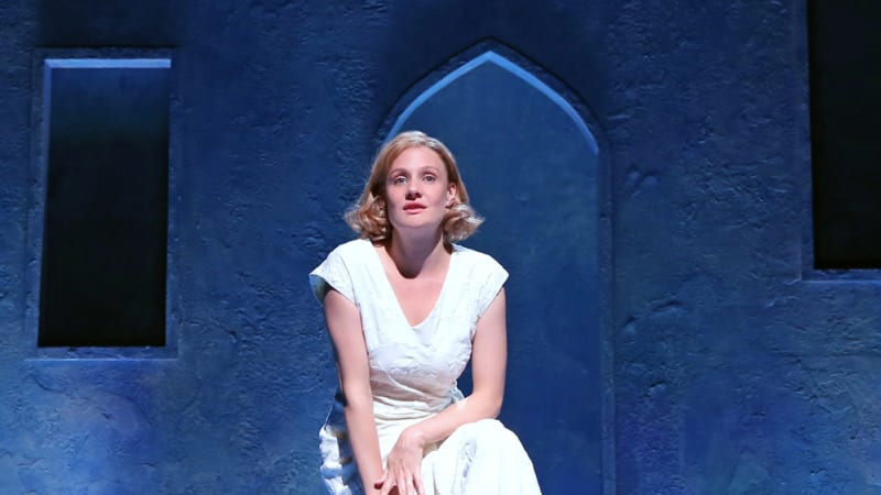 A blonde woman in a white dress sits on top of her suitcase under blue lighting, white petals scattered across the floor.