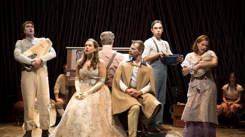 Around a man playing a piano is a young man holding a lap harp, a woman in a golden dress, a man in a cape, a man mixing a bowl, and a woman holding a baby.