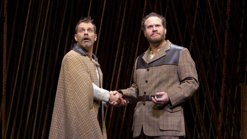 A man in a plaid cape shakes hands with a man in a plaid suit.