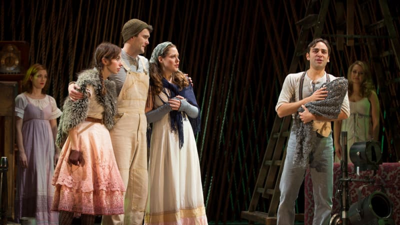 A man holding a baby in a blanket sings while a woman in a furry cape, a man in overalls, and a woman in peasant clothes watch him.