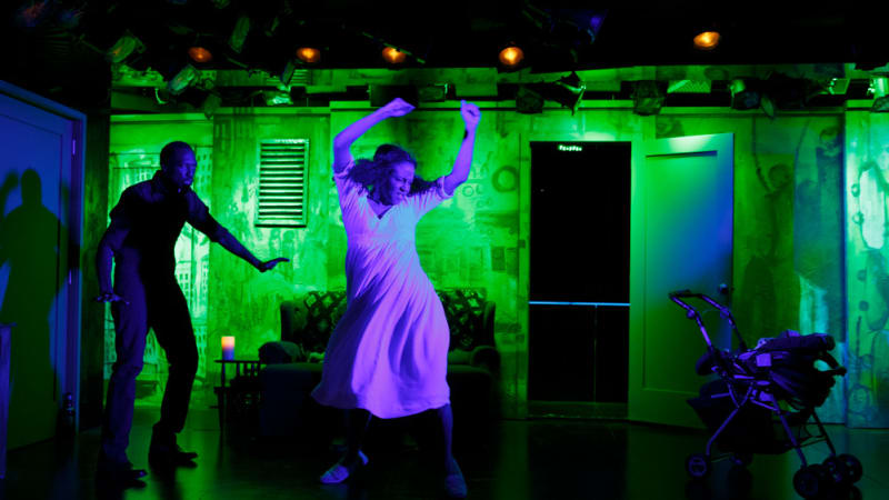 A woman in a white dress dances under green and black-lighting, her dress glowing purple. A man stands behind her cautiously.