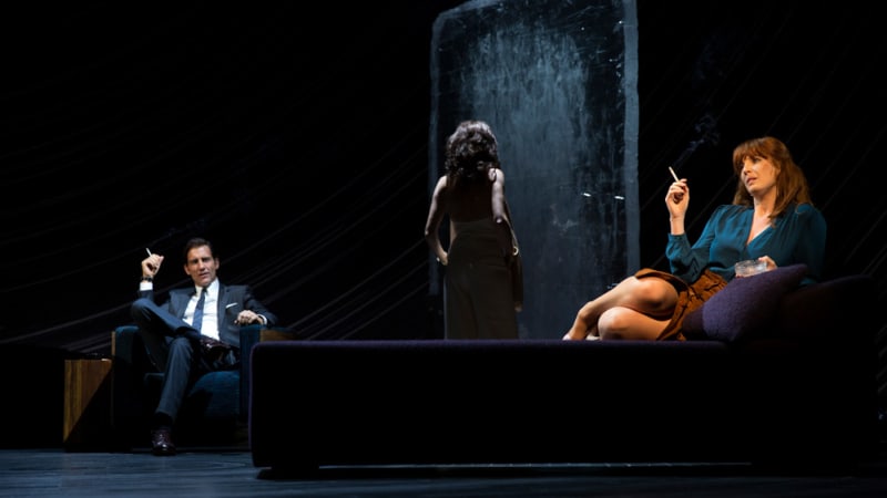 A man in a suit sits in a blue chair with a cigarette in his hand. A centerstage woman in a white dress is silhouetted. A woman in a blue blouse with a cigarette lounges on a purple bed.