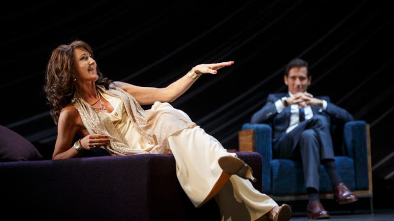 A woman in a silky white dress leans on her elbow on a dark purple bed telling a story. A man in a suit sits in a blue chair with his legs cross and hands clasped, listening to her.