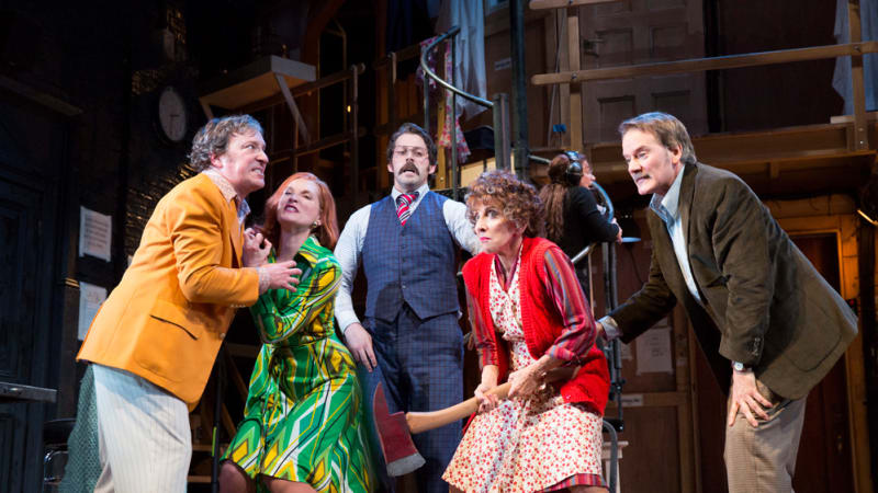 A clump of angry, eclectic actors. An older woman holds a fireman's axe, a man in yellow leans towards her but is held back by a woman in green, and two men look on at the action.