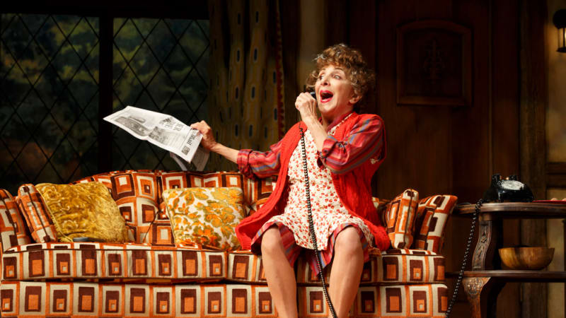 An older woman dressed in red sits on a brightly patterned couch. She excitedly speaks into a telephone and holds a newspaper in her other hand.