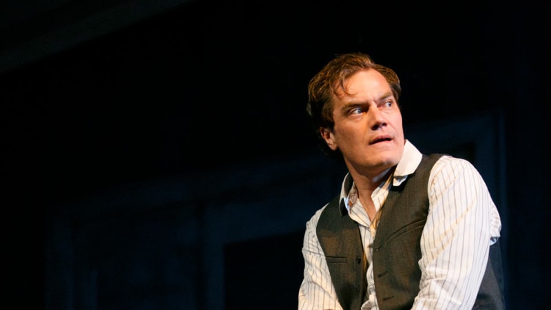 A man in a vest and collared shirt with a concerned expression leans against a wooden table. Behind him, another man in a vest sits in a low chair.