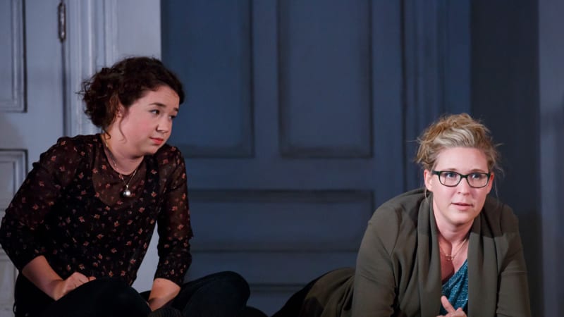 Two women sit on a wooden floor in a hallway. One lays on her stomach and rests on her elbows, and the other sits cross legged with her hands in her lap.