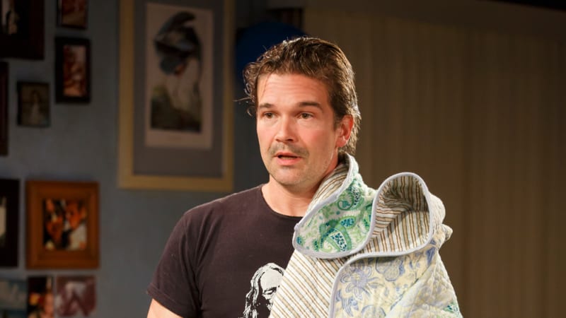 A man in a graphic tee stands by a softa, half draped in a quilt. He holds a small radio. On the coffee table next to him is a six pack of beer.