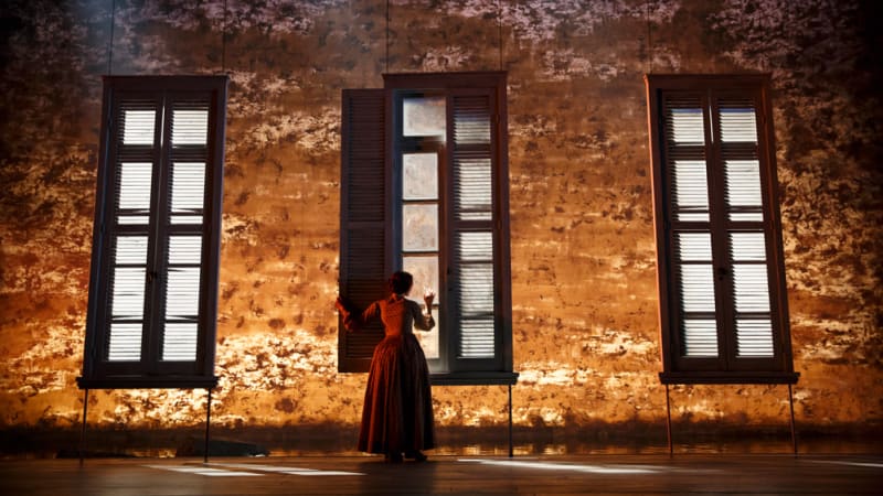 A woman alone on stage in an 1860s dress opens the shutter of a giant window and holds a hand to the glass. The background looks like gold and purple crushed velvet.
