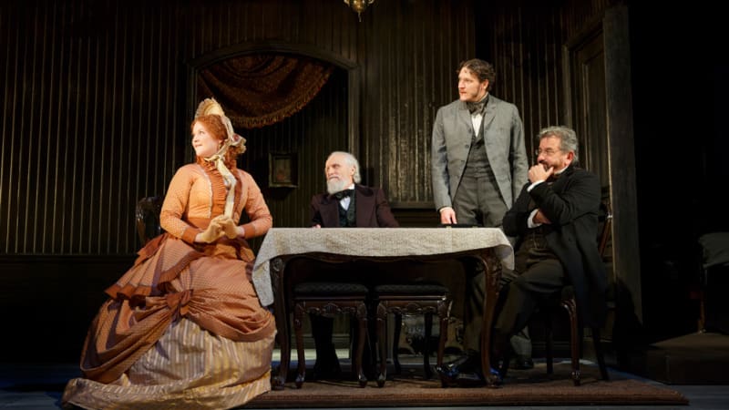 A group of upperclass 1860s-styled people sit and stand around a small table in a drawing room. Three men of varying ages in suits. One woman in an intricate orange dress and bonnet.
