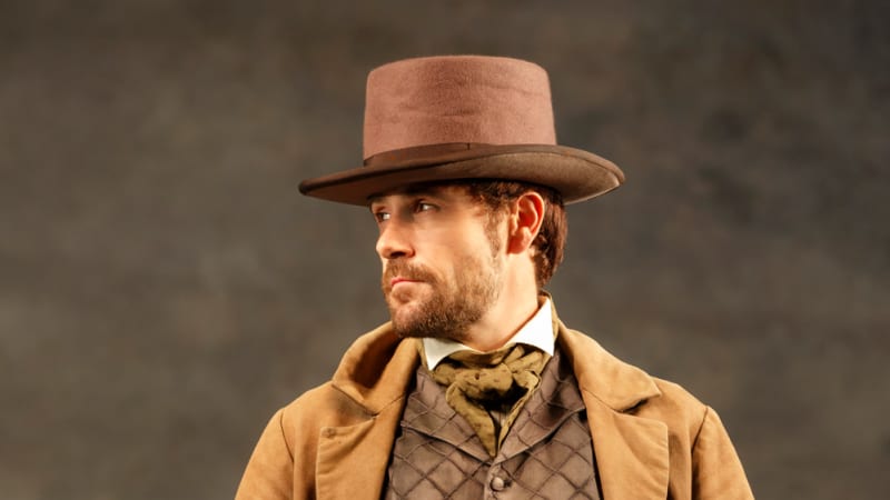 A man in an 1860s brown suit, long coat, and top hat stands with his hands in his pockets.