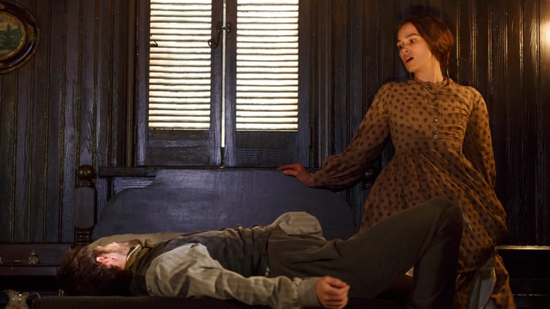 A man lays unconscious on a wooden bed. A woman in a brown dress stands against the wall with one hand on the headboard, she's in shock.