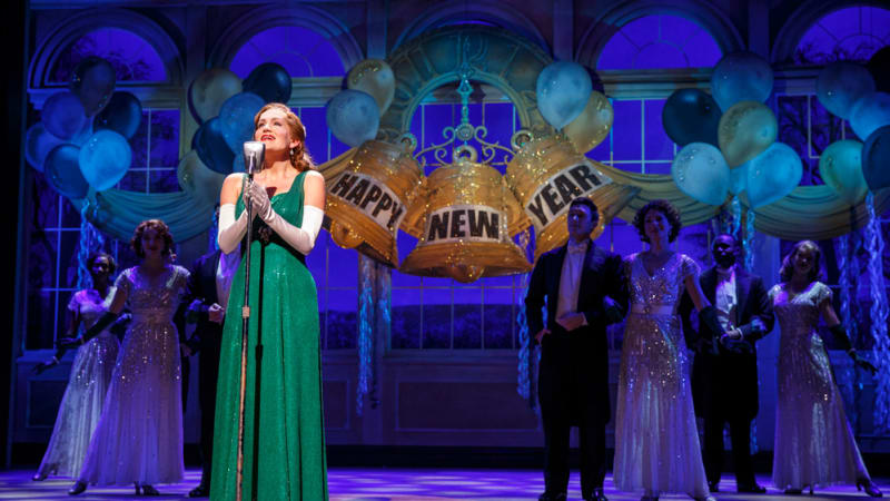 A woman in a shimmering green dress with white satin gloves sings at a microphone at a fancy New Year's Eve party.