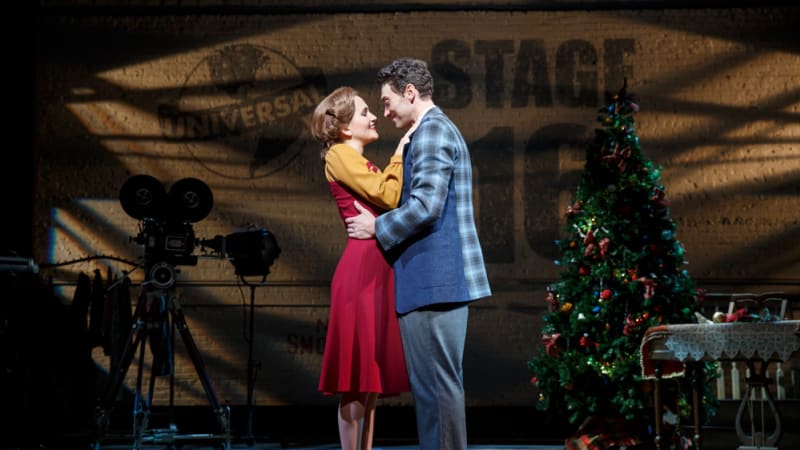 A woman in a dark red and yellow dress places her hands on the chest of a man wearing a blue checkered blazer. He holds her waist in a romantic moment behind a soundstage wall.