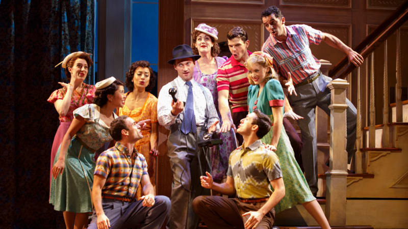 A group of people in bright 1940s style clothes excitedly gather around a staircase and surround a man in a navy fedora and tie holding a telephone.