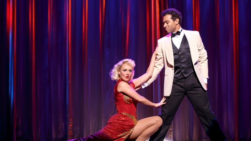 A woman in a red and gold sparkly dress lunges down dramatically next to a man in a tux and rests a hand on his leg and his arm.