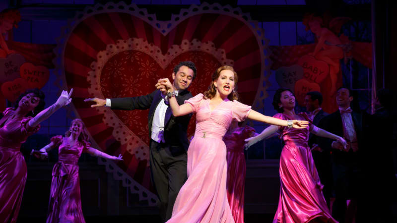 A man in a tux dances with a woman in a long pink dress. Other tux and dress duos dance around them. The backdrop behind them is full of lacy hearts.