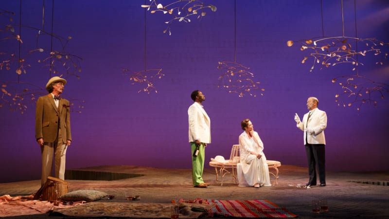 Abstract, wispy cherry tree branches float over a barren stage with a purple and blue gradient background. Picnic blankets and a garden bench lay on the stage. Four actors stand and sit around.