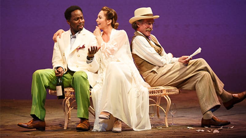 A woman in white sits on a garden bench between two men. One wears green pants, a white blazer, and holds a bottle of wine. The other wears a white hat and tan suit set while holding a paper.