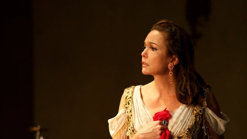 A woman in a golden corset and red patterned skirt sits on a chair and holds a handkerchief to her chest with sorrow.