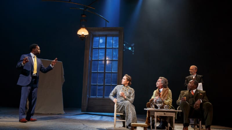 A group of fancy people sit at small chairs at a small table in an empty room with one large window and a hanging light. A man off to the side in a suit speaks to them and gestures.