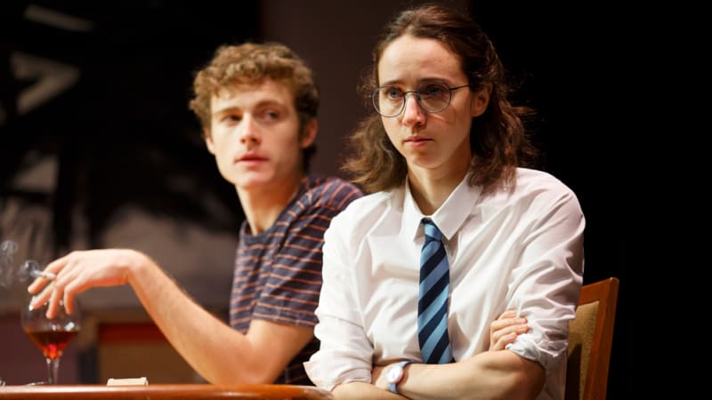 A young man and woman at a kitchen table. The woman has glasses and wears a school uniform. She crosses her arms. The man has curly hair and a striped shirt. He holds a cigarette and has a wine glass.