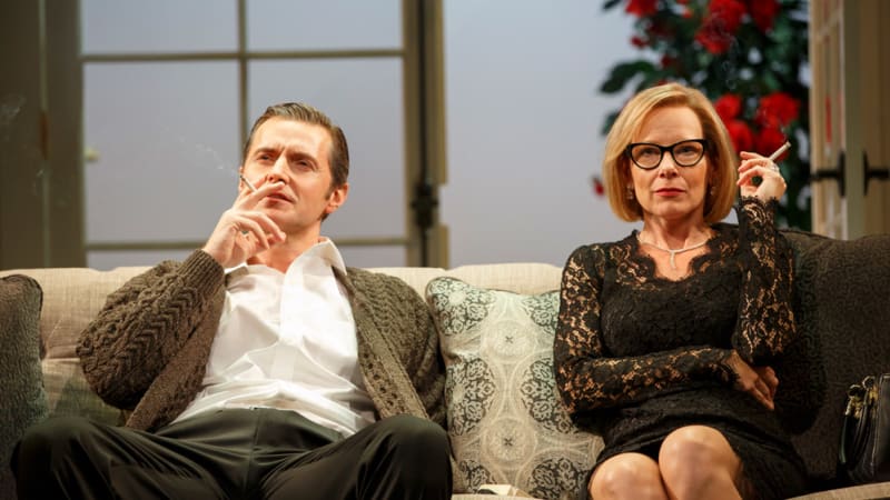 An older man and woman sit together on a sofa, each holding a cigarette. The man wears a cardigan over a collared shirt. The woman wears a black dress made of lace, glasses, and a pearl necklace.