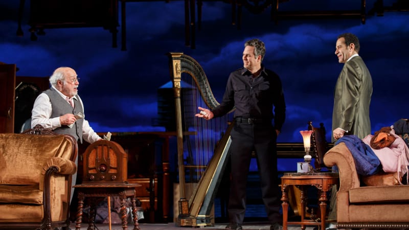An old man in glasses and a business suit holds money in his hands and talks to a man in a police uniform and another in a green suit. Around them is antique furniture, including a harp.