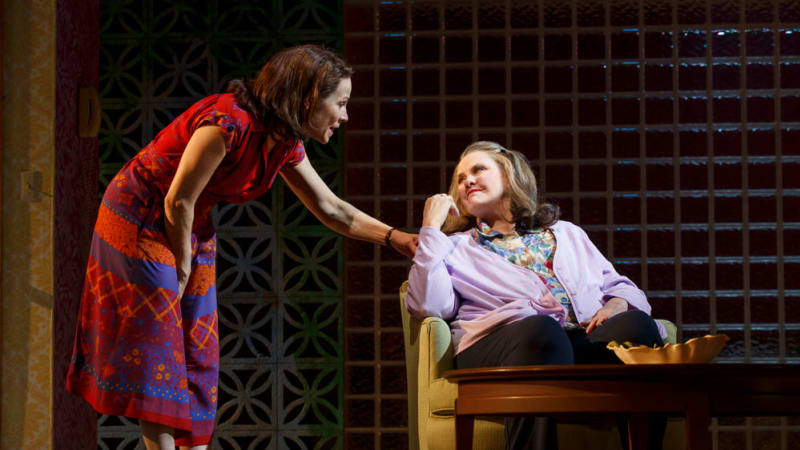 A woman in a red and blue patterned dress leans down and places a hand on the shoulder of a woman in a purple cardigan and floral shirt, who sits in a motel room chair.