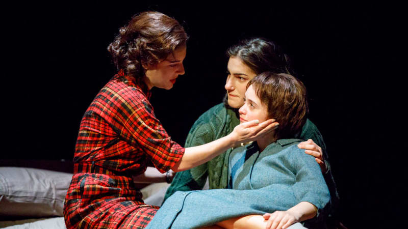 Three young women in their pajamas sit together on a bed, the youngest being held by one while the other rests a hand on the youngest's cheek.
