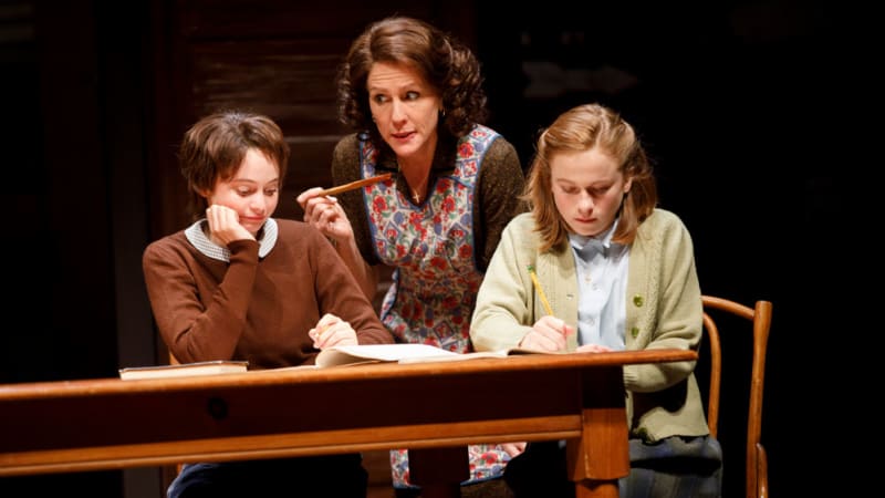 Two young woman sit at a wooden table doing schoolwork, while an older woman in an apron leans in between them.