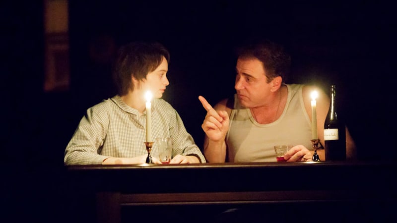 A young woman sits next to an older man at a table, illuminated only by two tall candles. He points a finger at her while holding a glass of liquor in his other hand.