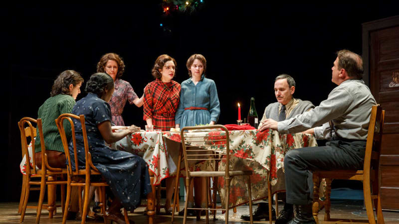 A family sits at a long dining table with multiple tablecloths and mismatched chairs. The energy is tense. Three of the women stand up near the end of the table.